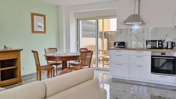 A modern kitchen with white cabinets, a built-in oven, and a coffee maker. There is a dining area with a wooden table and chairs, and a sofa in the living room.