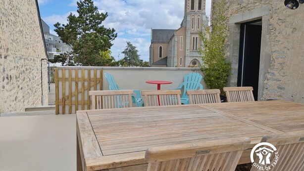 An outdoor patio with a wooden table and chairs, a small red table, and a view of a church tower. - Modern cottage in Louverné, sleeps 6-8, pets allowed, parking (Louverné)