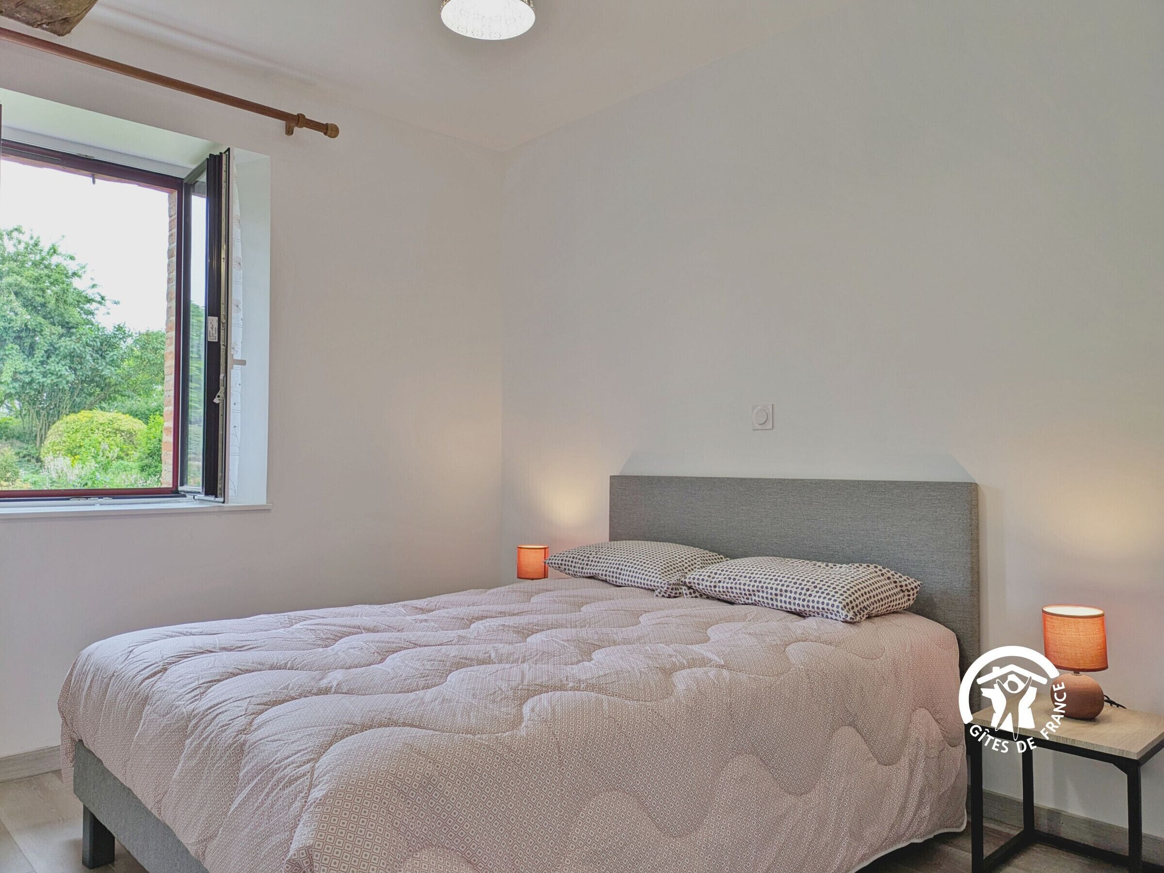 A bedroom with a bed, a nightstand, a lamp, and a window with a view of greenery.