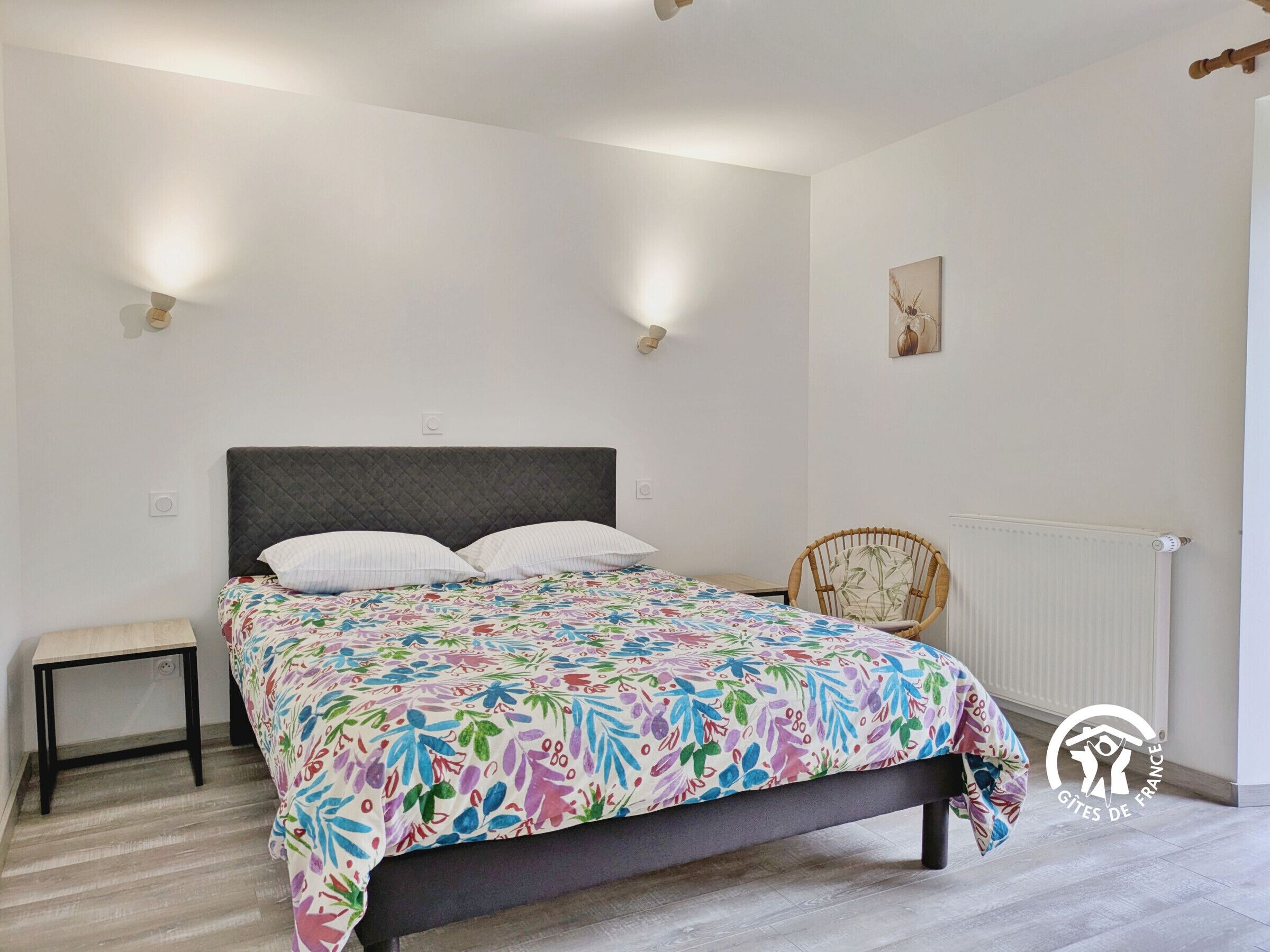 Ein Schlafzimmer mit einem Bett, einem Nachttisch und einem Stuhl. An den Wänden sind Lampen und ein Heizkörper angebracht.