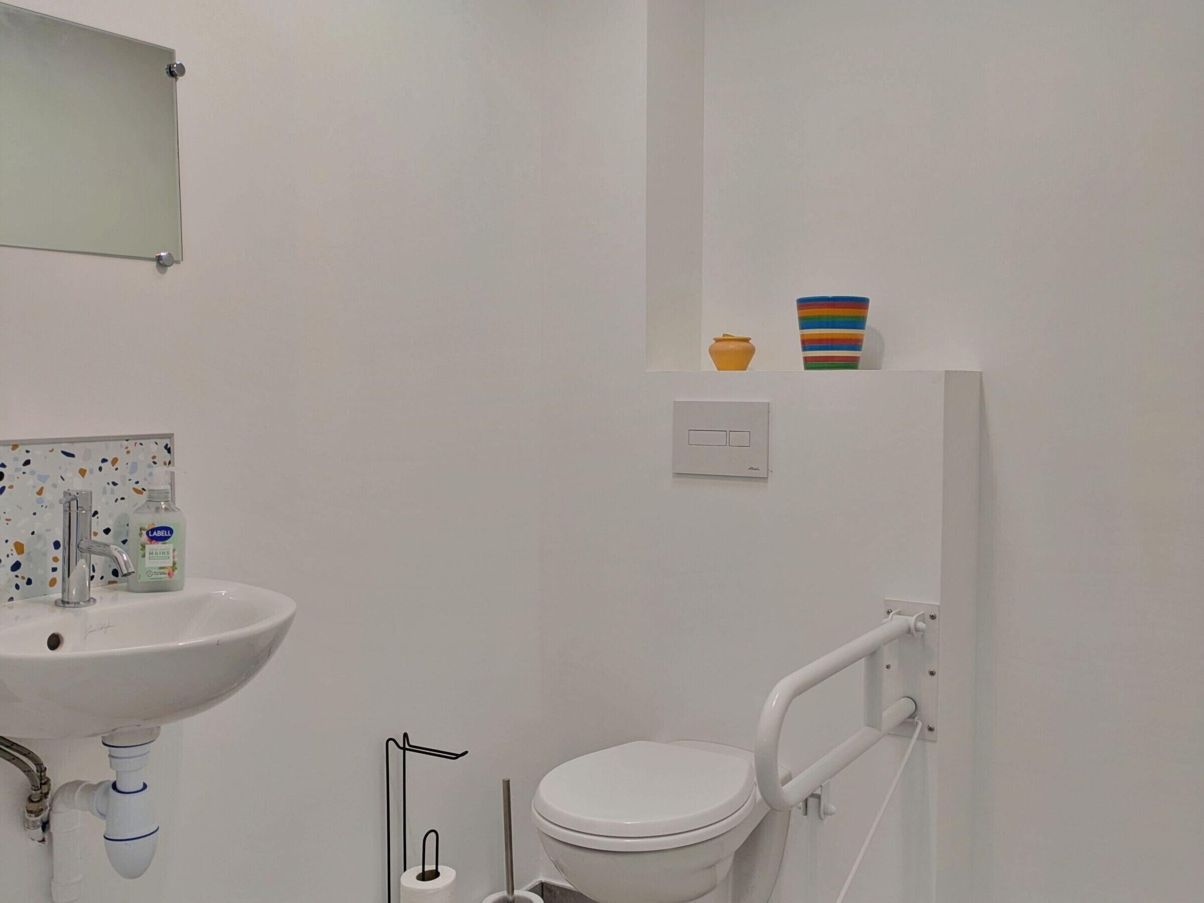 A bathroom with a white sink, a mirror, a toilet, and a small shelf with a vase and a striped container.