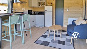 Une cuisine compacte avec des meubles blancs, une cuisinière noire et un réfrigérateur. On y trouve un coin repas avec deux chaises et une petite table. Un canapé bleu et un tapis à carreaux se trouvent dans l’espace salon.