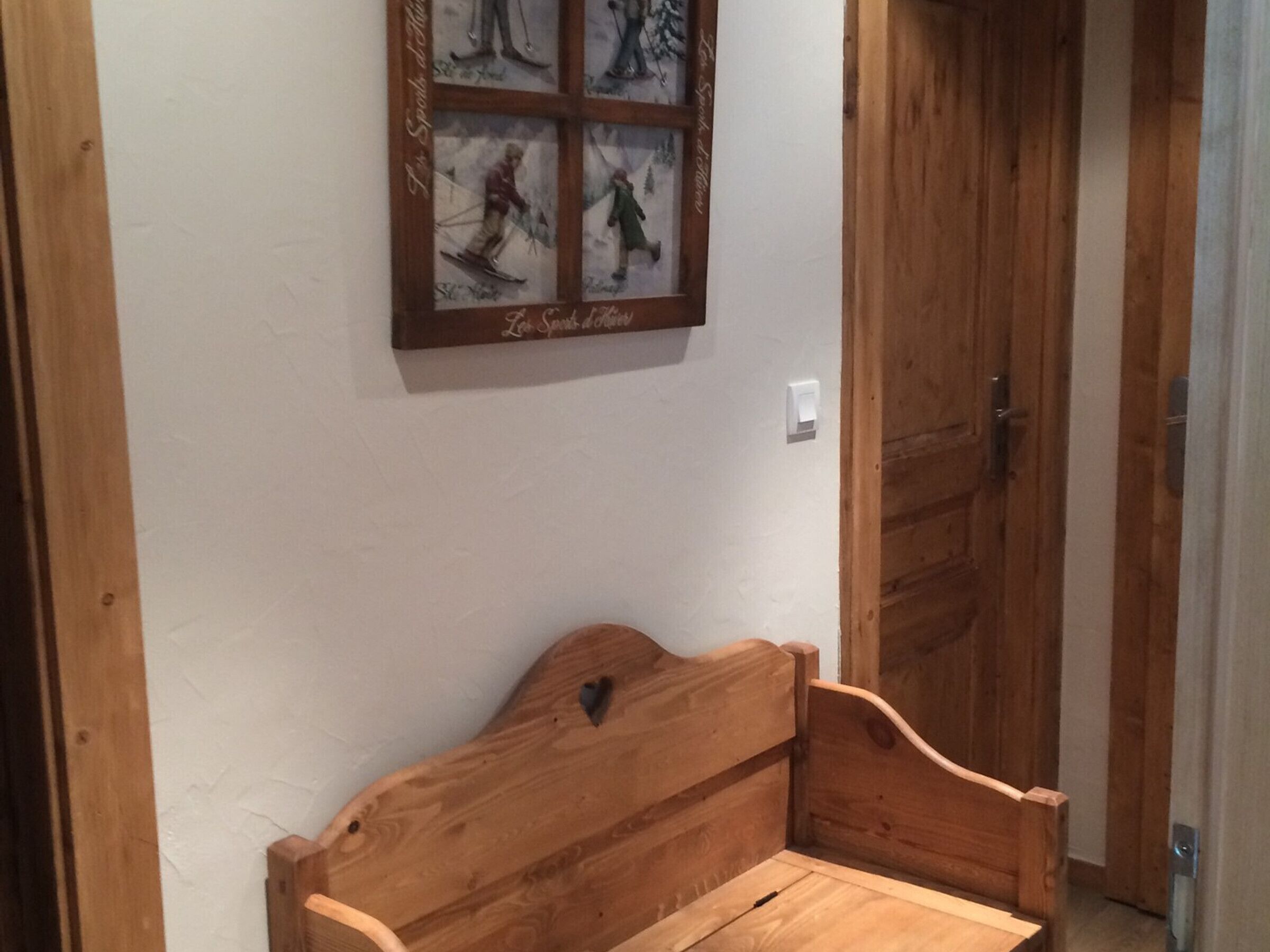 Un banco de madera con un recorte en forma de corazón en el respaldo, ubicado en un pasillo con puertas de madera y una fotografía en el marco de esquiadores en la pared.