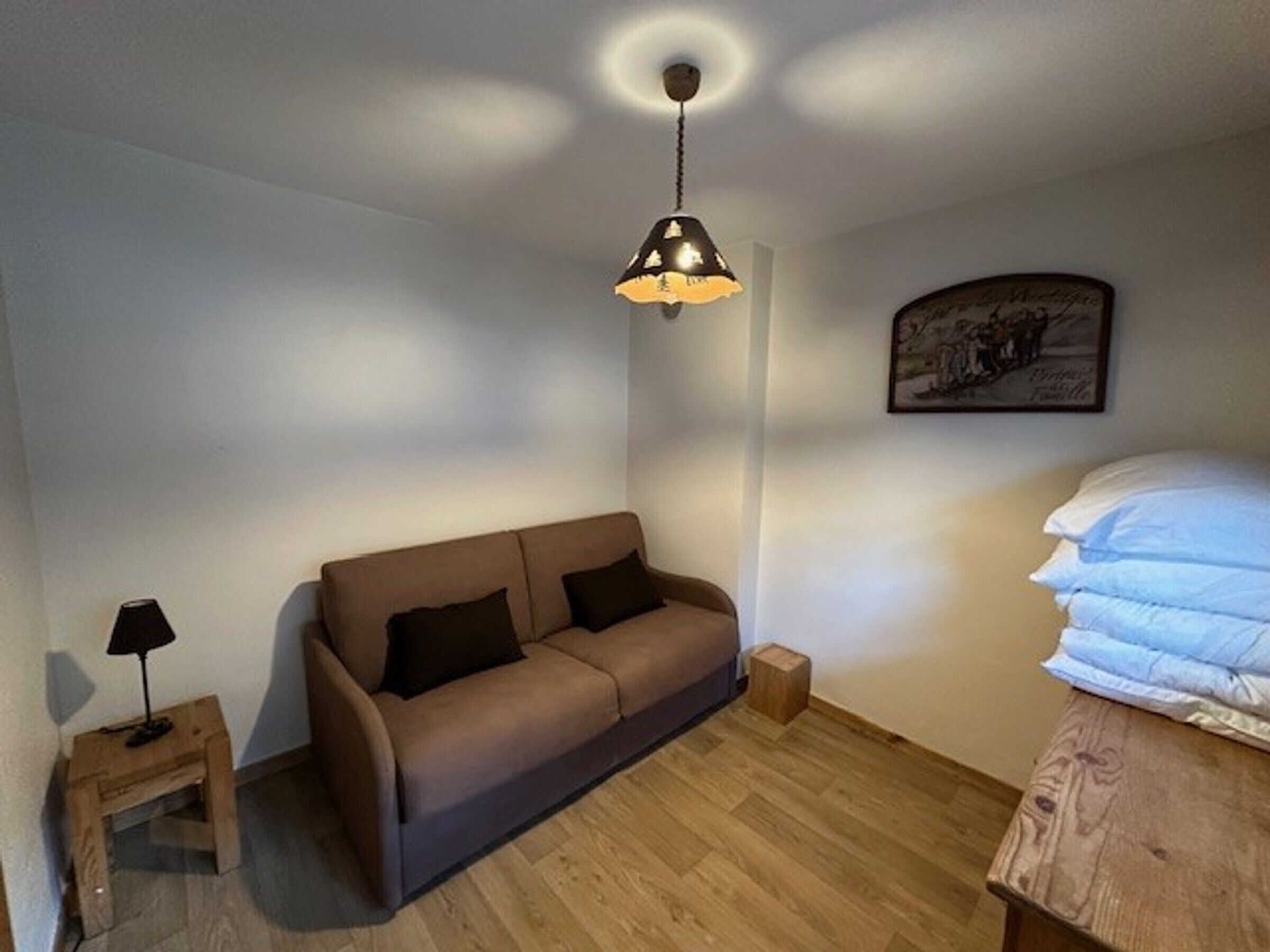 A small, cozy room with a brown sofa, a small wooden side table, a lamp, a bed with white bedding, and a ceiling light fixture.