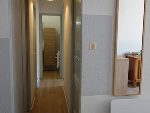 A narrow hallway with a wooden floor, a mirror on the right, and a door at the end leading to a room with a bed and a small table.
