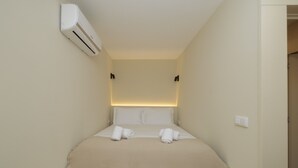 A small, neatly made bed with a beige bedspread and two rolled towels, a wall-mounted air conditioner, and a light switch on the wall.