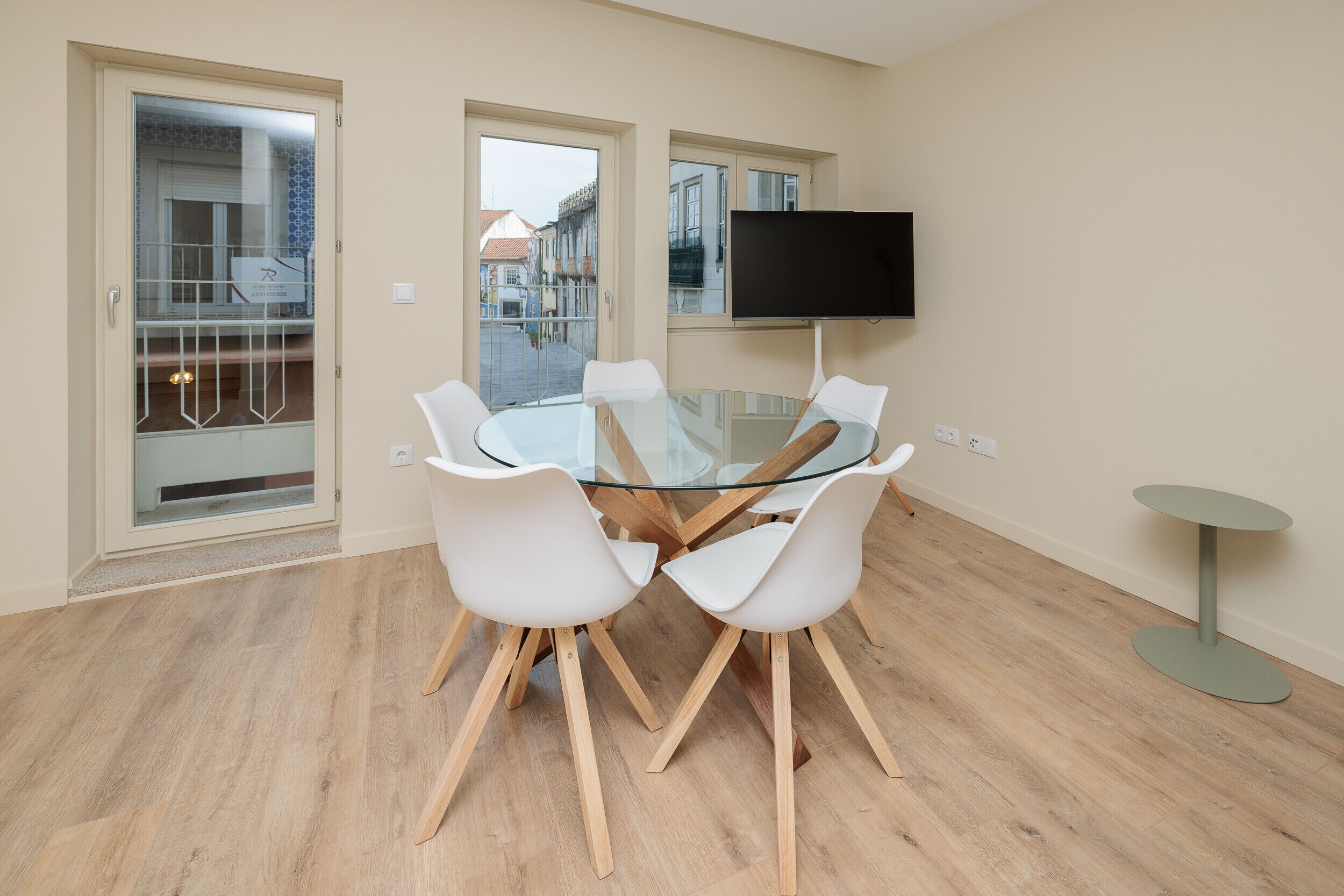 Una sala moderna con una mesa de vidrio para comer rodeada de sillas blancas, un televisor montado y una pequeña mesita auxiliar.