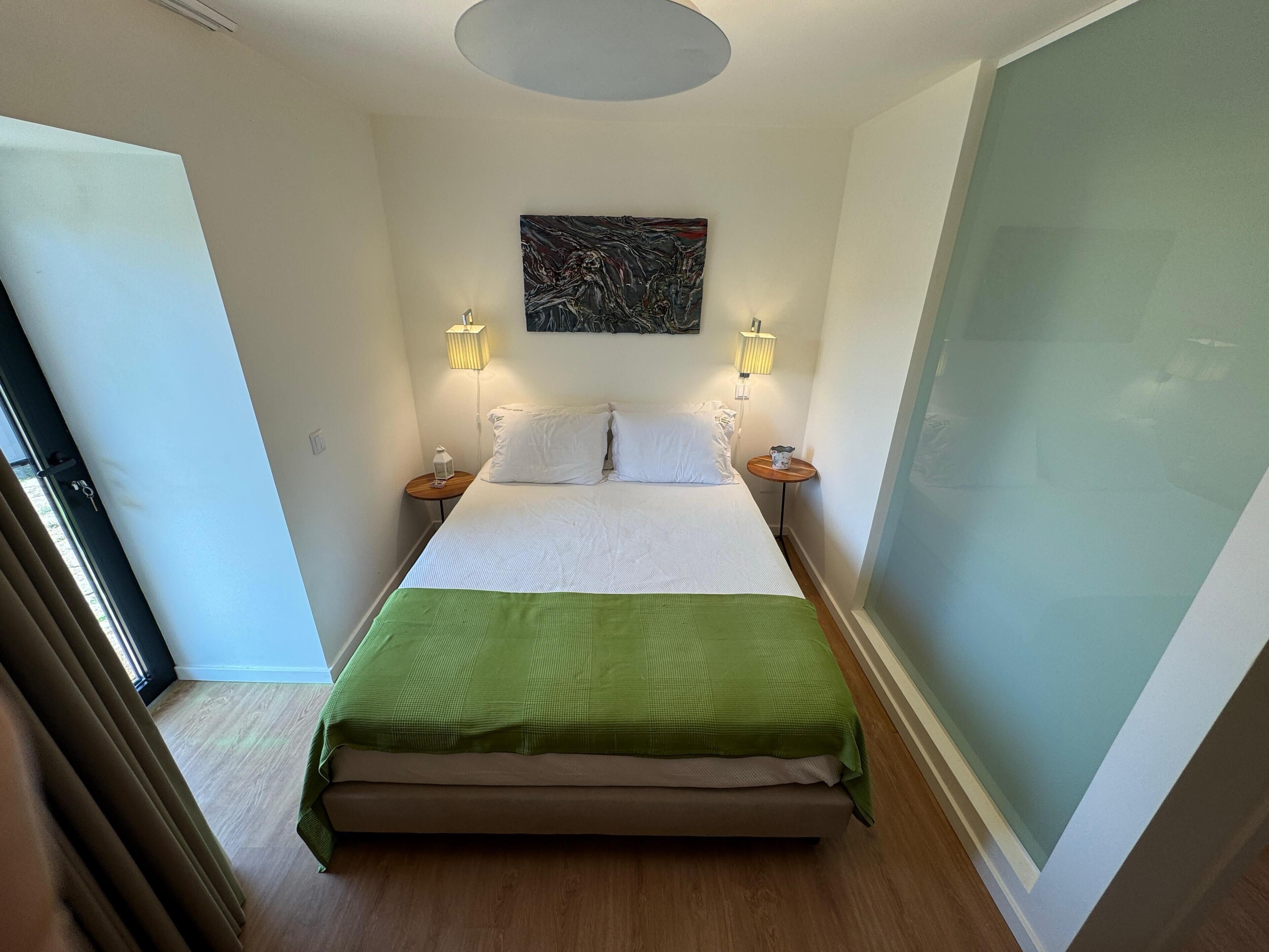 Habitación de hotel con cama, dos lámparas de noche, una manta verde y un cuadro en la pared.