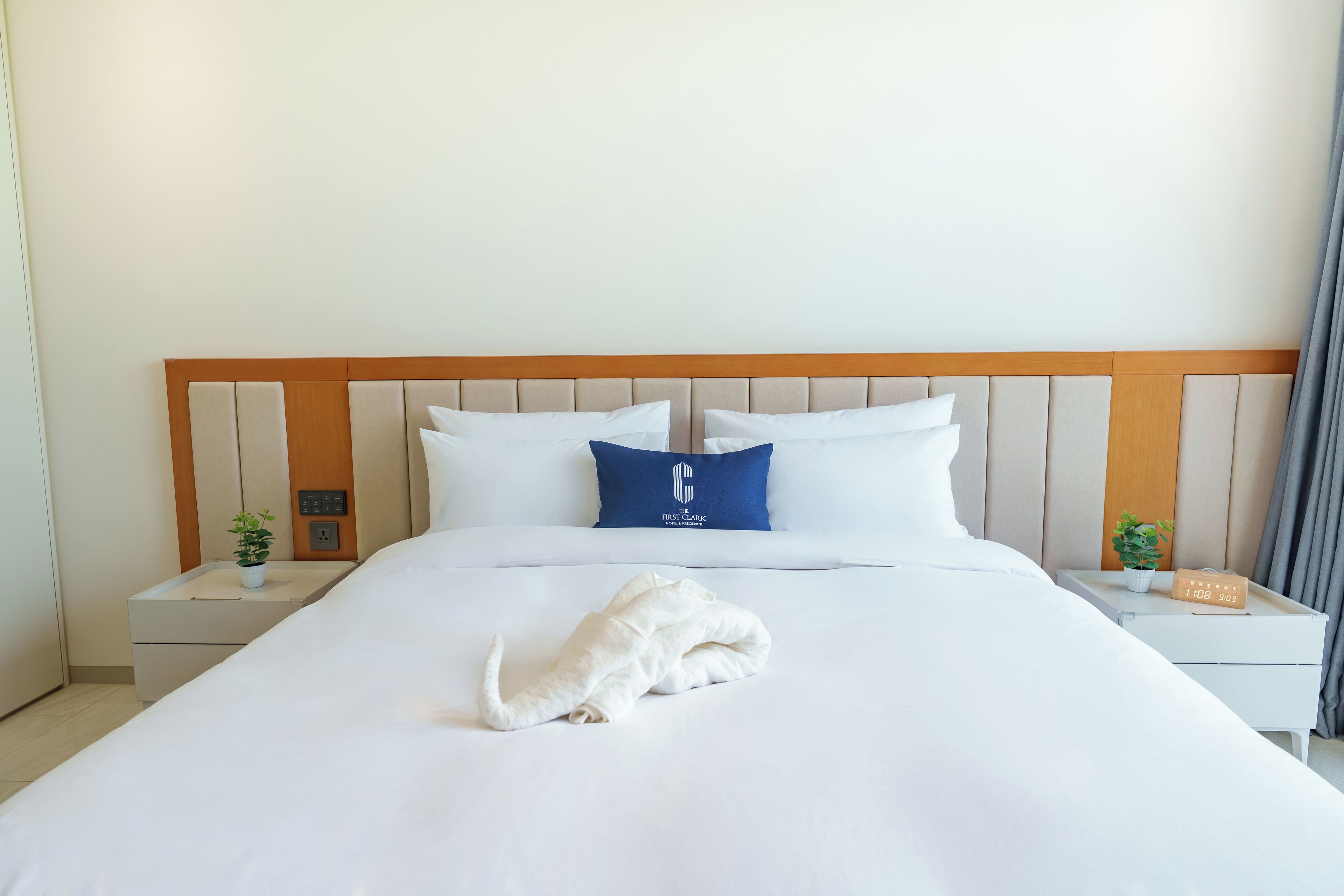 A neatly made bed with a white blanket, a blue pillow, and a white towel folded on the bed. There are two bedside tables with small potted plants and a digital clock.