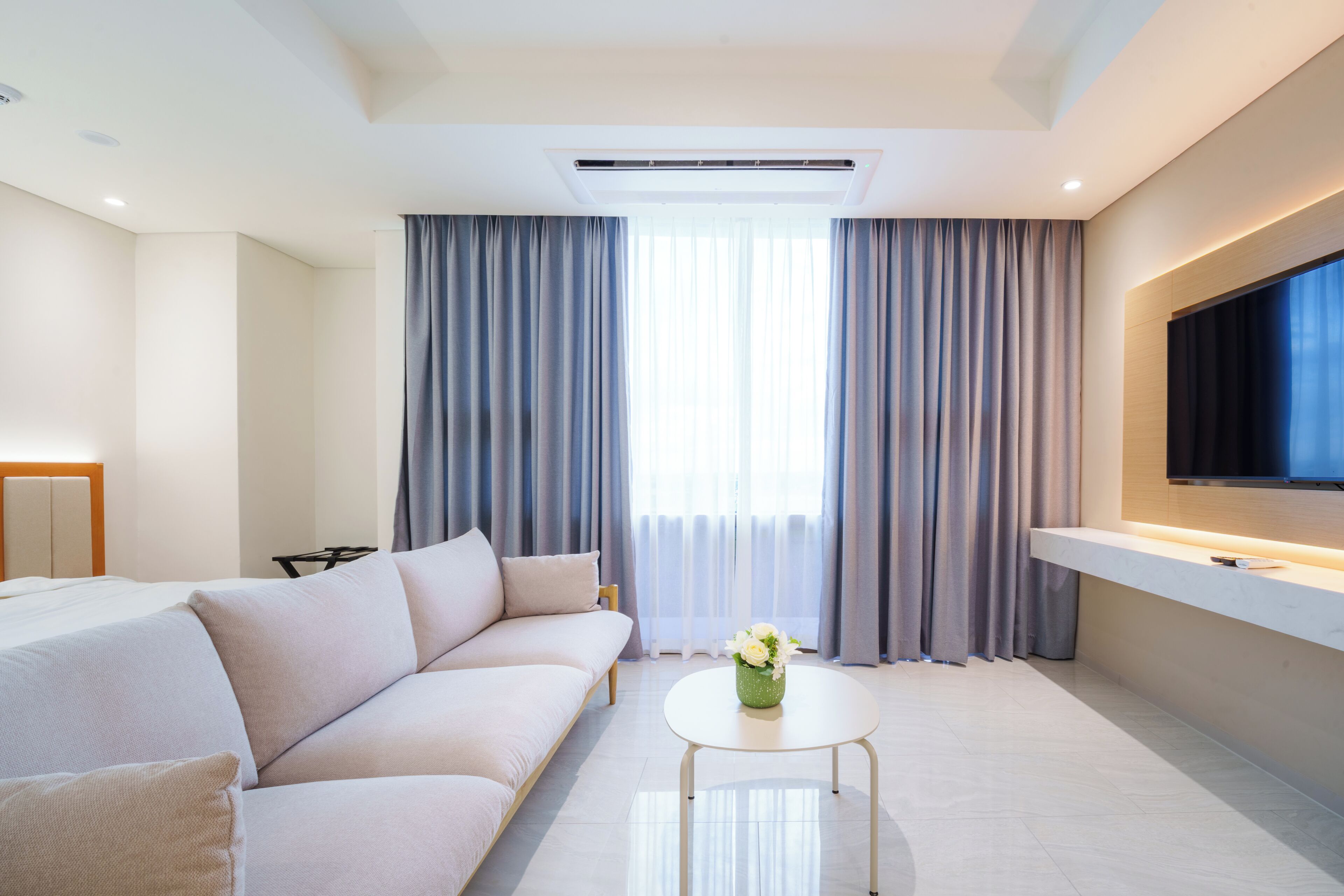 Une chambre d’hôtel moderne, dotée d’un canapé, d’une petite table ronde, d’un téléviseur à écran plat fixé au mur et d’une grande fenêtre avec des rideaux.