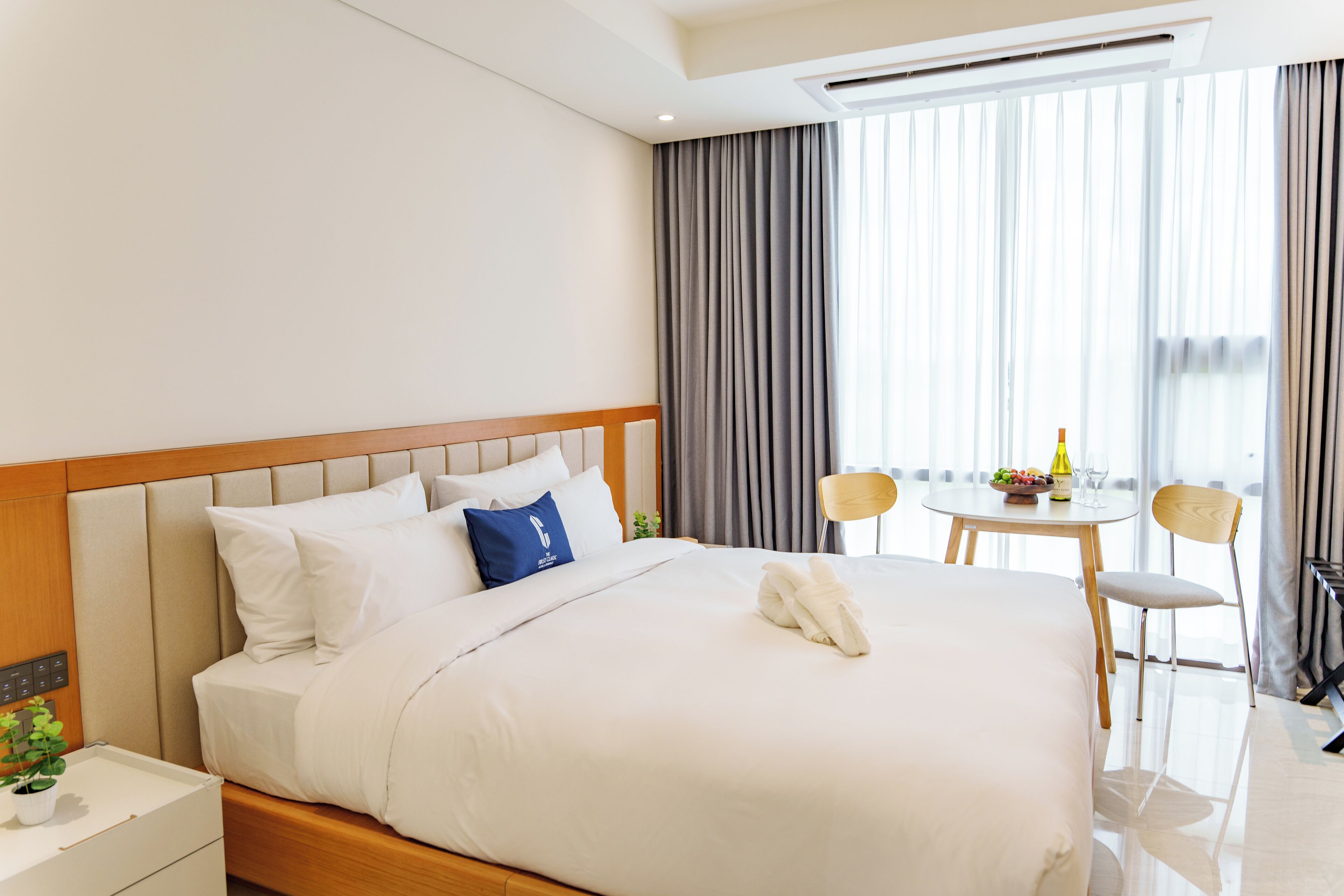 A hotel room with a neatly made bed, a small table with a bottle and fruit, and a chair.