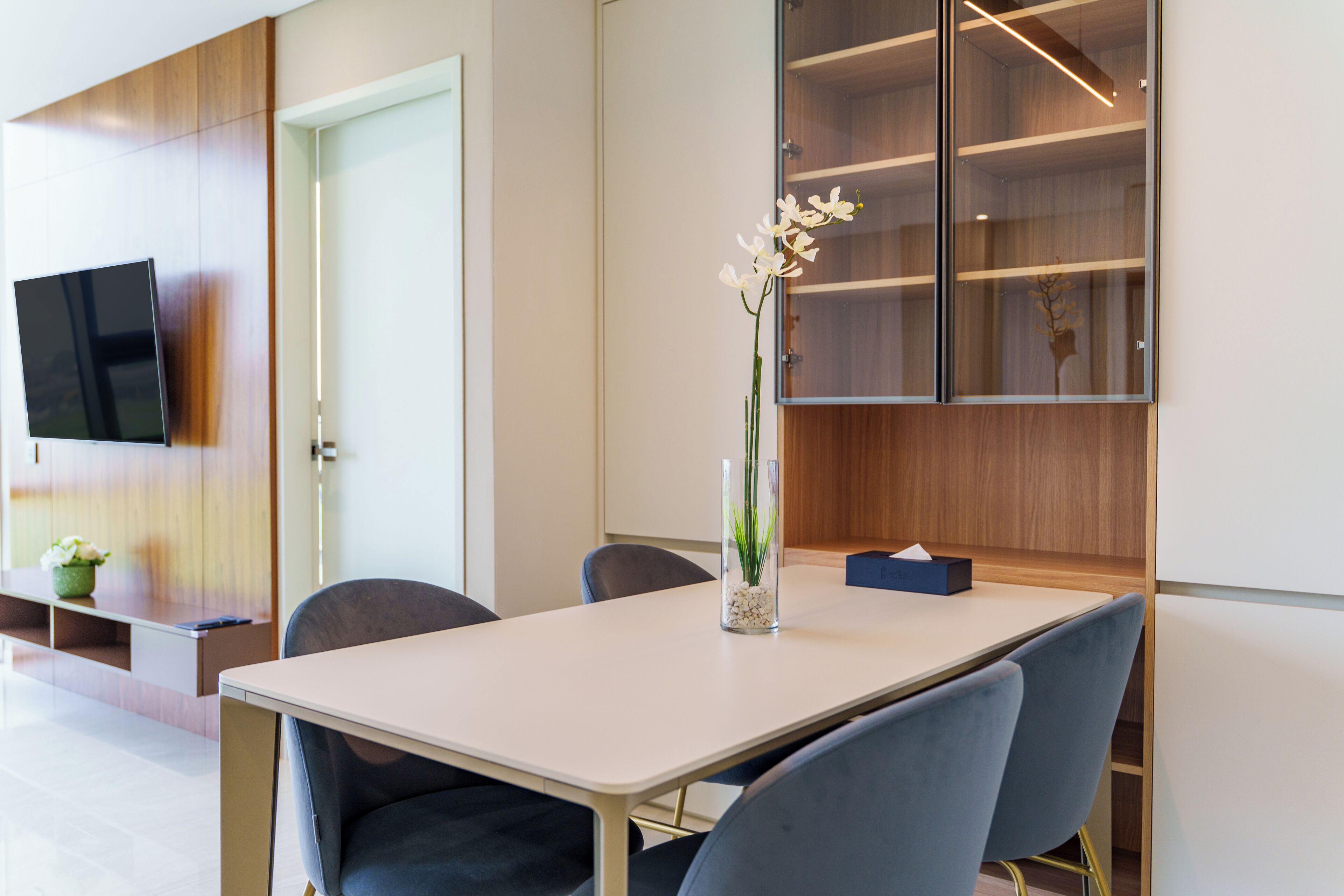 A modern conference room with a white table, blue chairs, a vase with white flowers, a flat-screen TV on the wall, and wooden paneling.