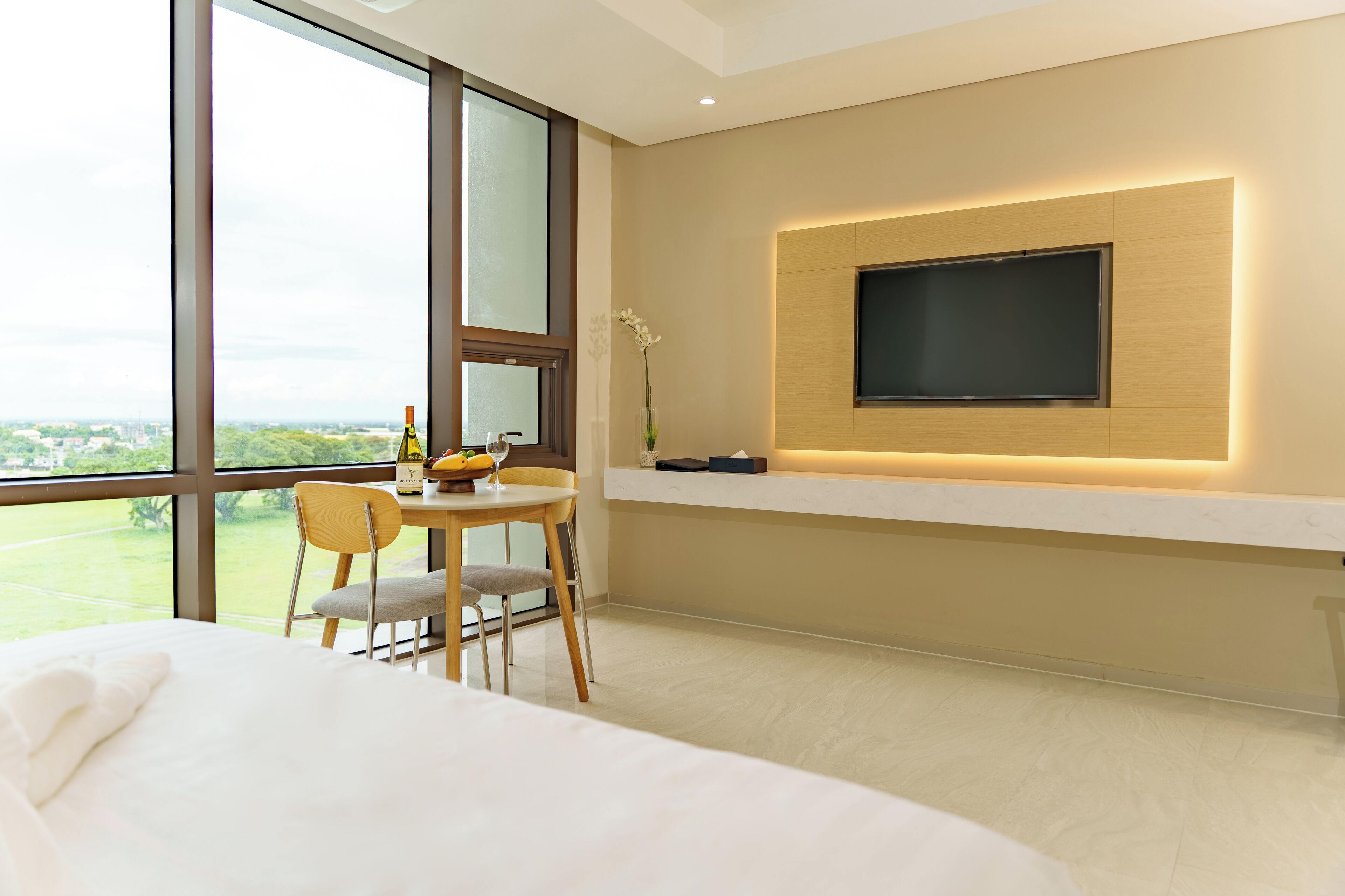 A modern hotel room with a large window, a bed, a dining area with a table and chairs, and a mounted TV on the wall.