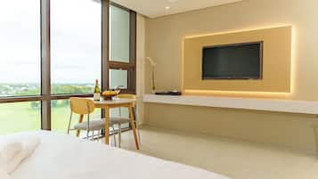 A modern hotel room with a large window, a bed, a dining area with a table and chairs, and a mounted TV on the wall.