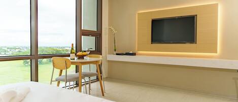 A modern hotel room with a large window, a bed, a dining area with a table and chairs, and a mounted TV on the wall.