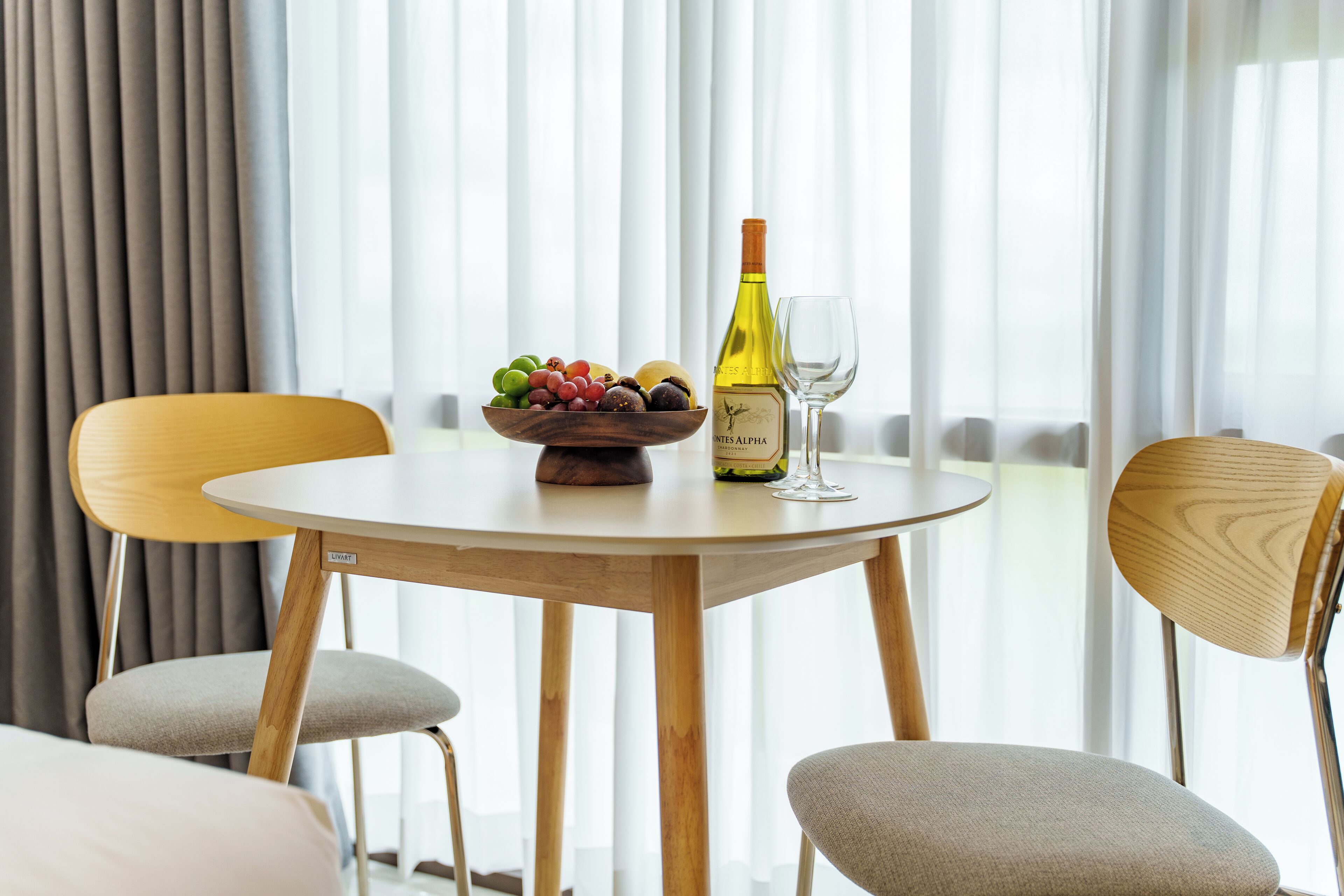 A round wooden table with a bowl of fruit, a bottle of wine, and two wine glasses.