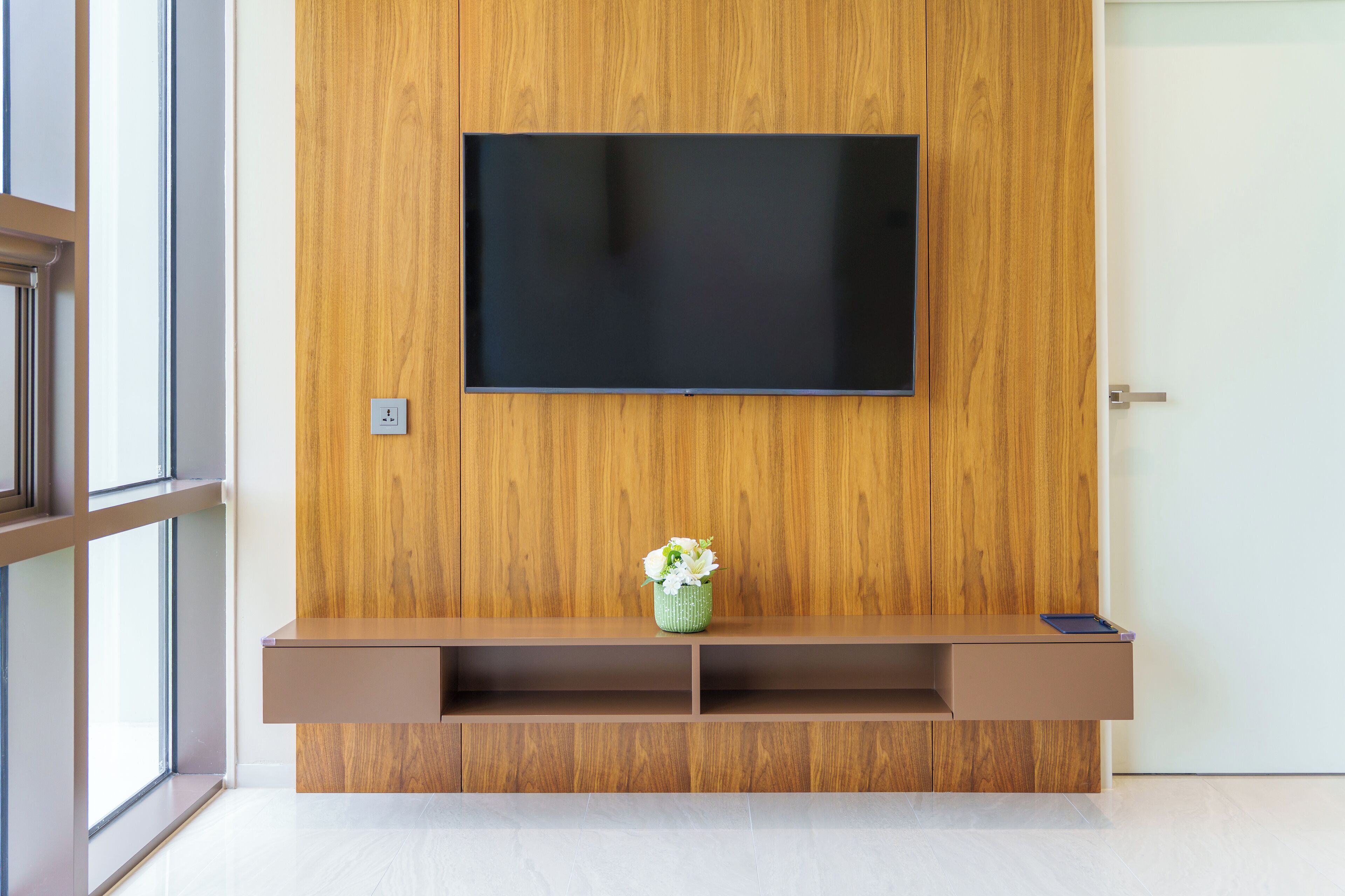 A modern living room with a wooden wall, a mounted TV, a small shelf with a vase, and a door.