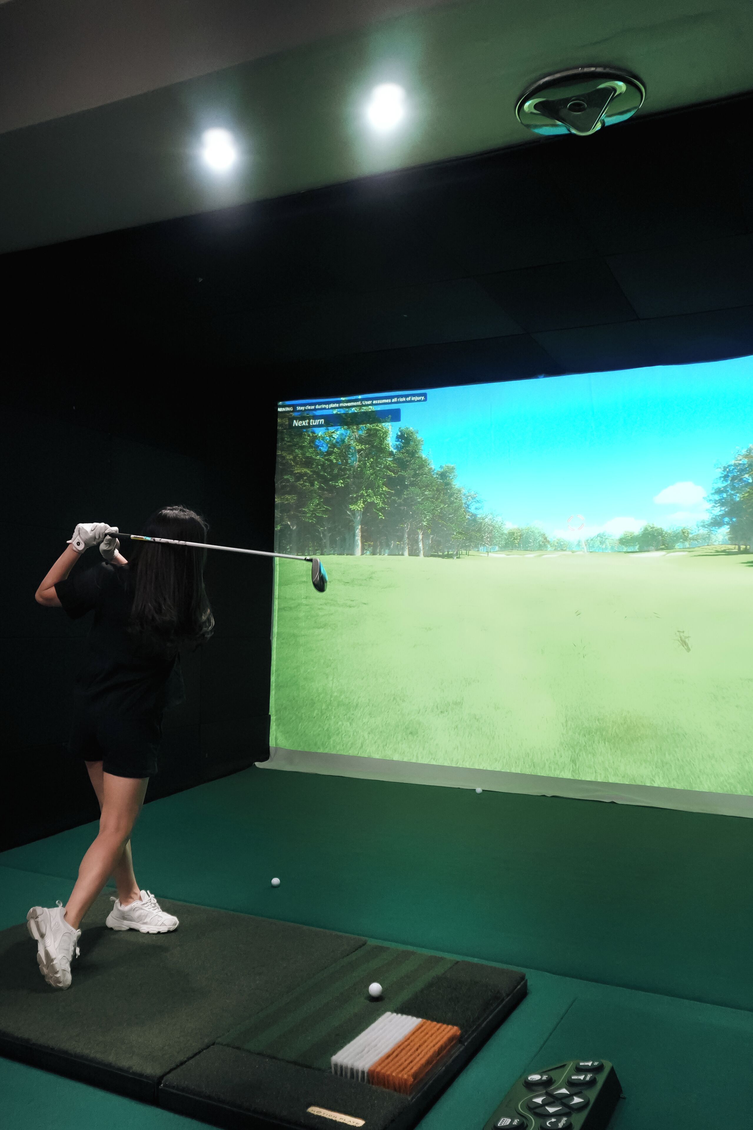 Une personne qui pratique le golf à l’intérieur, devant un écran qui affiche un parcours de golf.