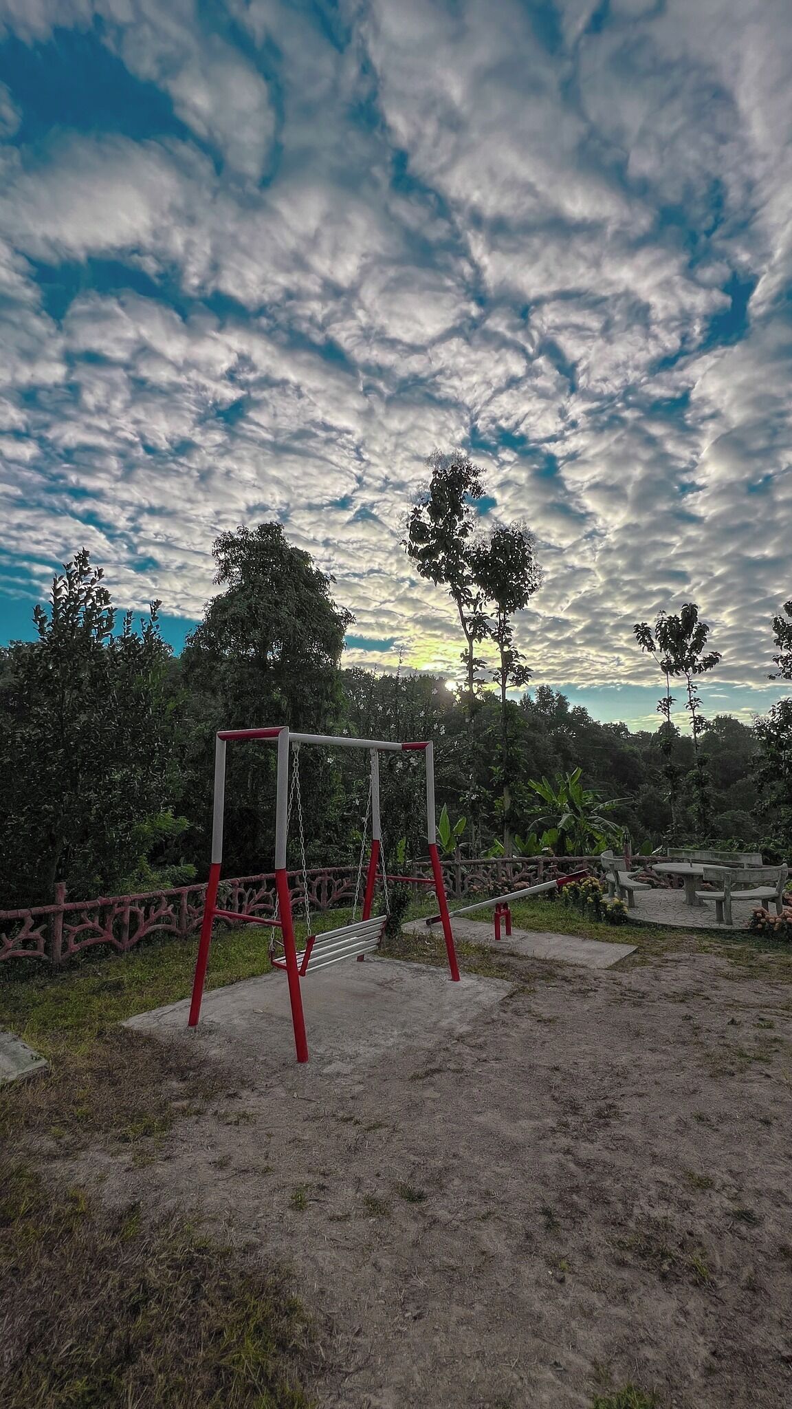 Um parquinho com balanços e escorregador, cercado por árvores e uma grade.