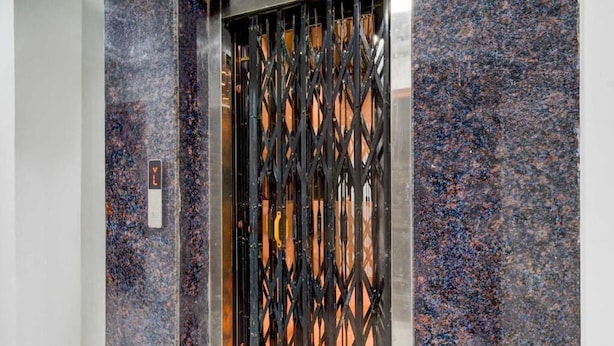 Un ascensor moderno con una puerta metálica geométrica y un muro de mármol.