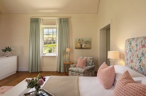 A well-lit room with a bed, a floral armchair, a side table with a lamp, and a painting on the wall.