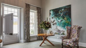A spacious room with a high ceiling, a large window, a chandelier, a floral armchair, and a large abstract painting. - Brookdale Estate - Manor House (Cape Town)