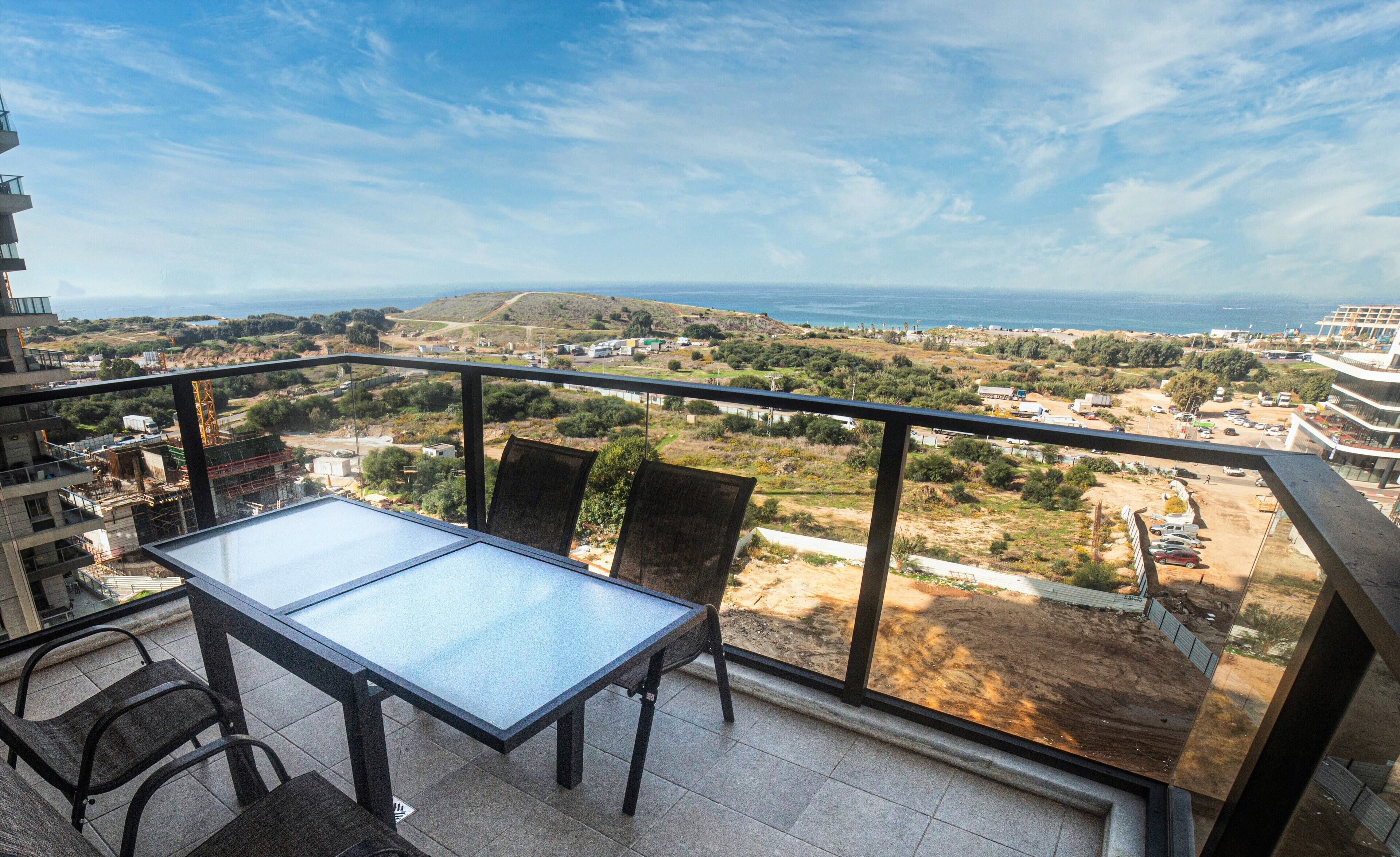 A balcony with a table and chairs, overlooking a coastal landscape with buildings and construction.