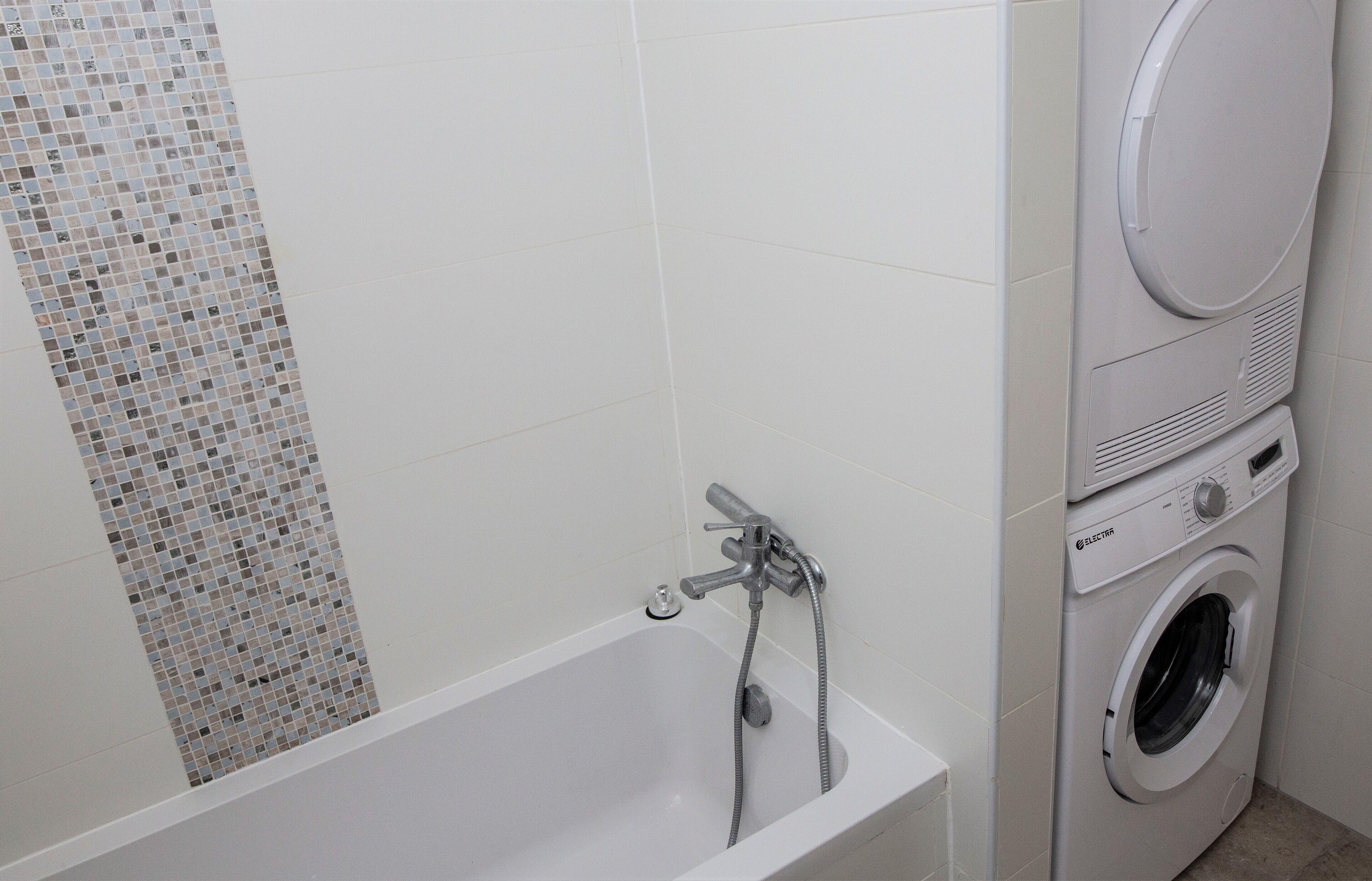 Une salle de bain avec un mur recouvert de carreaux blancs, un motif en mosaïque sur le côté gauche, une baignoire blanche avec un robinet et un lave-linge à chargement frontal.
