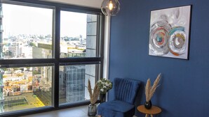 Una habitación moderna con un ventanal grande, una silla azul, una mesita redonda y un cuadro en la pared.