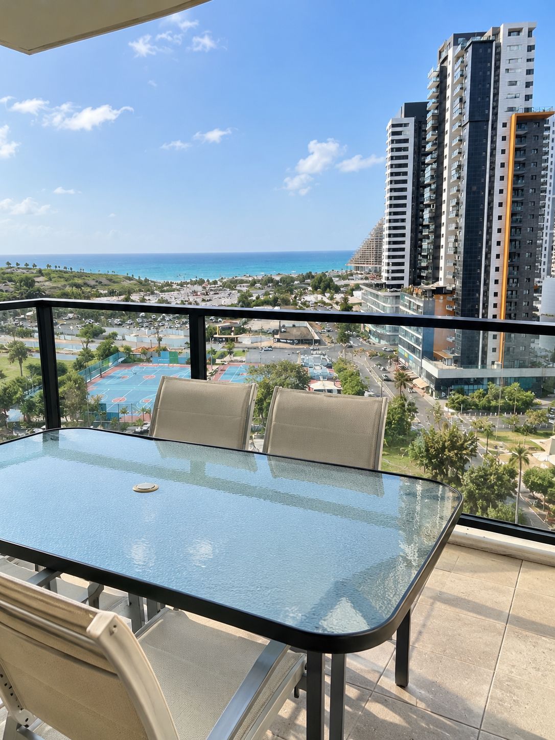 Un balcon doté d’une table et de chaises en verre, offrant une vue sur une zone côtière avec des bâtiments et un ciel dégagé.