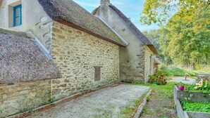 Ein Steinhaus mit Strohdach, einem kleinen Fenster und einem Garten mit Pflanzen und einer Bank.