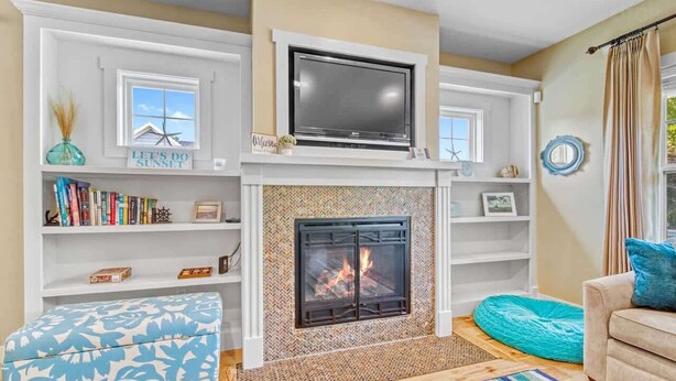A living room with a fireplace, a television mounted above it, and a bookshelf filled with books. - All In | Pool Access | Fire Pit | Game Room (South Haven)