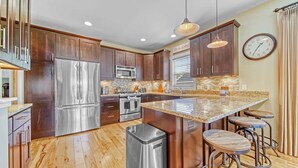 A modern kitchen with wooden cabinets, stainless steel appliances, a granite countertop, and a clock on the wall. - All In | Pool Access | Fire Pit | Game Room (South Haven)