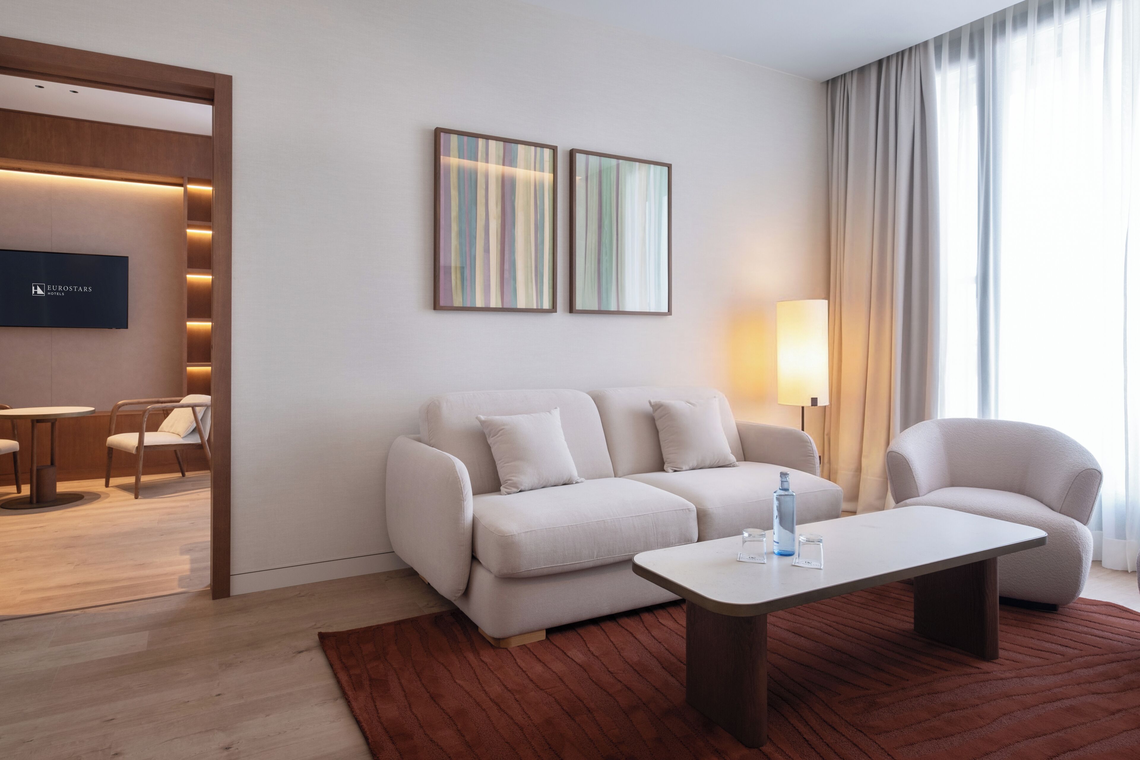 Una moderna camera d'hotel con divano, poltrone, tavolino, televisore e opere d'arte alle pareti.