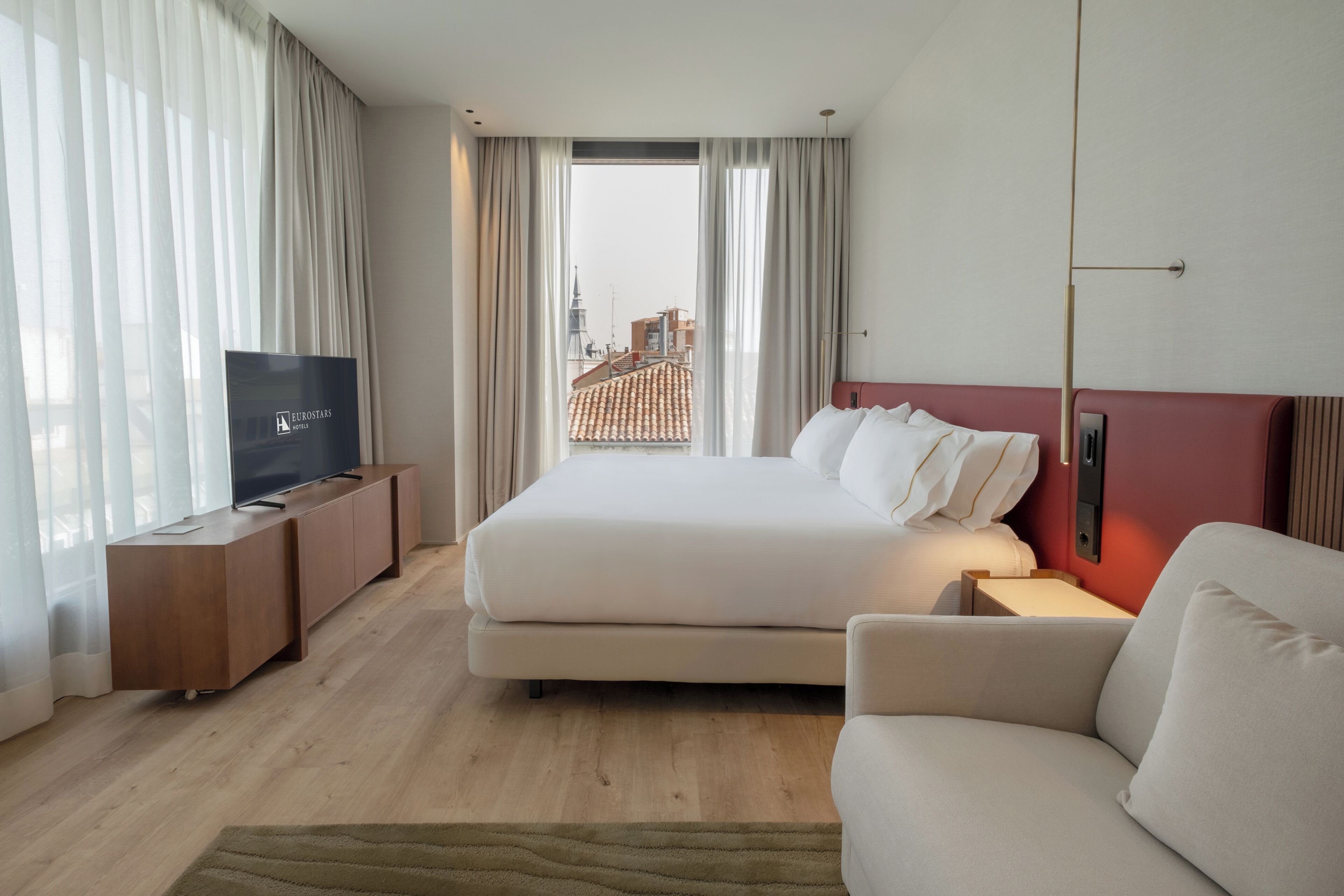Habitación de hotel con cama, televisión, sofá y vista a un paisaje urbano.