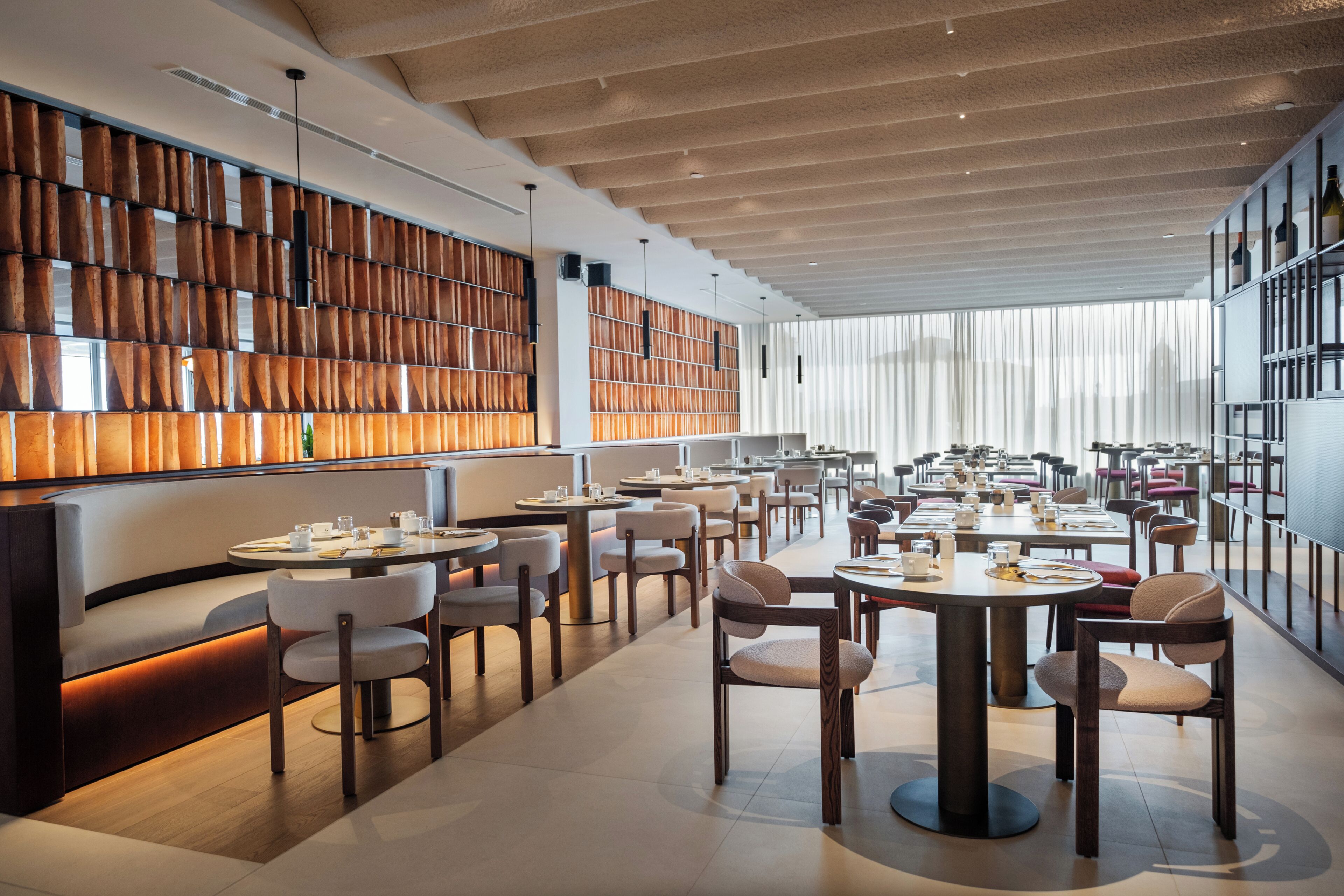 A modern restaurant with a long communal table, individual seating, and a wall of wooden panels.