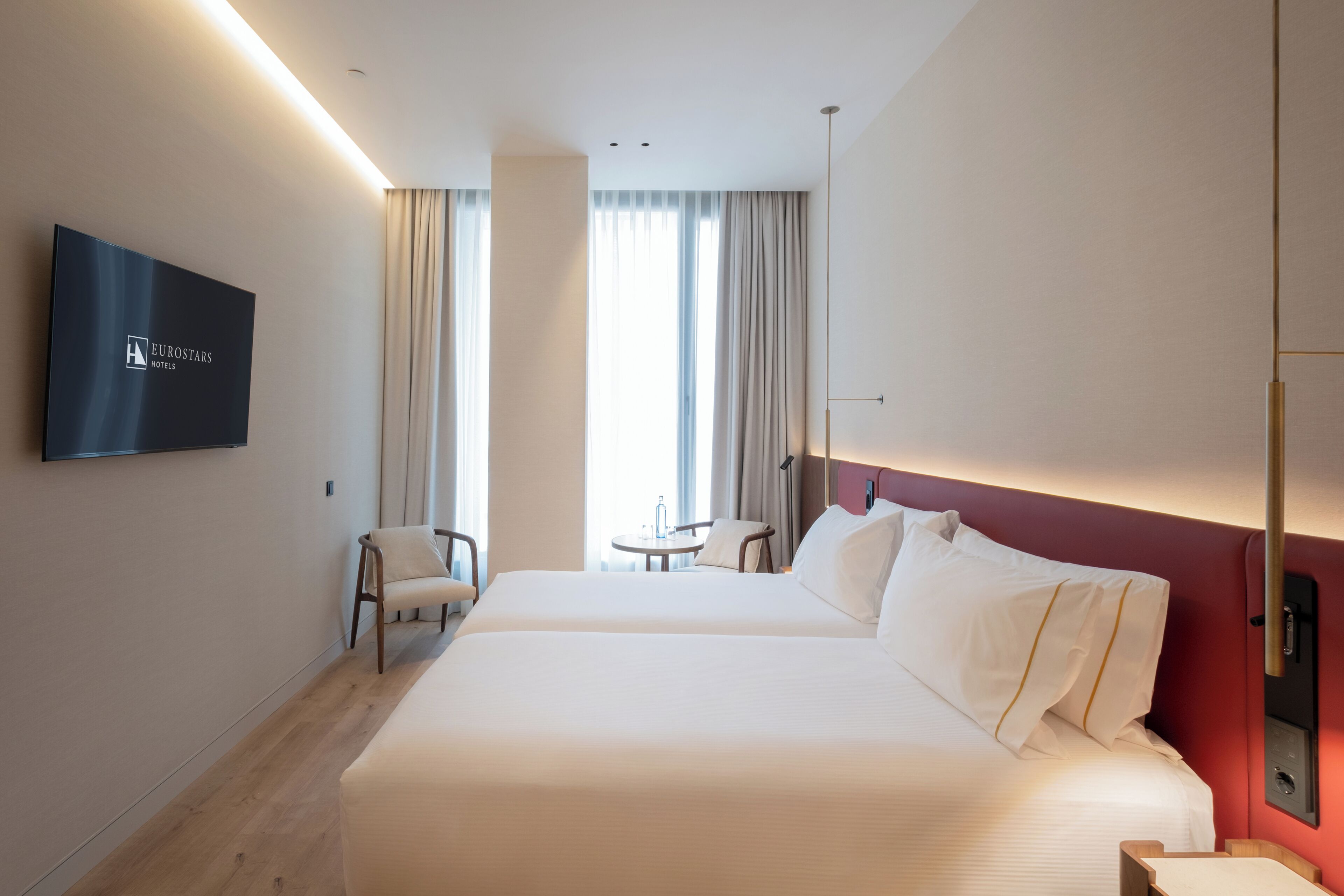 Habitación de hotel con cama, televisión, silla, mesita y vistas al exterior.