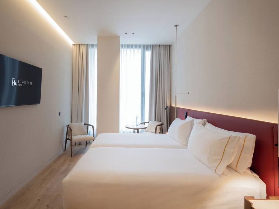 Habitación de hotel con cama, televisión, silla, mesita y vistas al exterior.