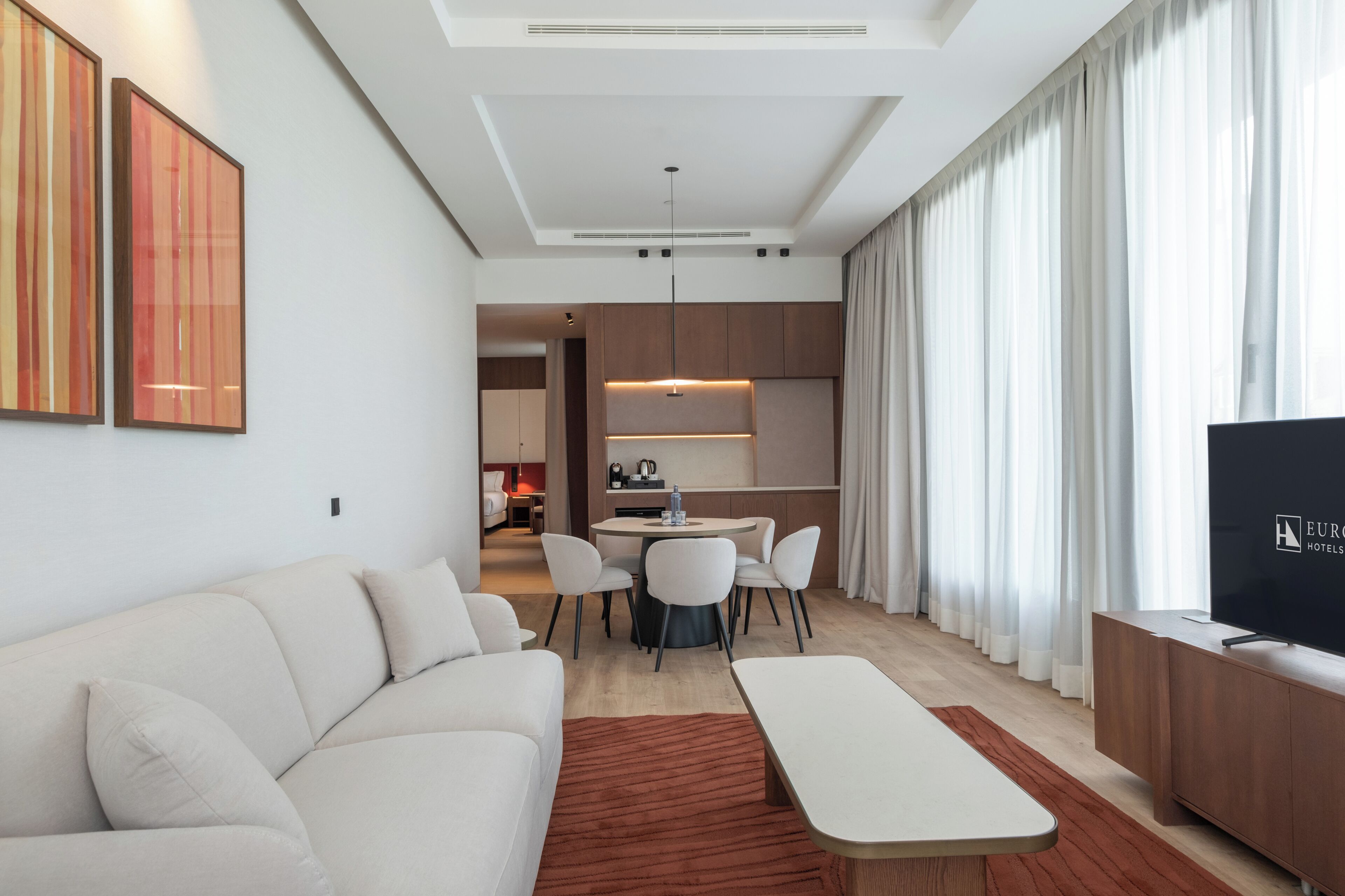 Una habitación de hotel moderna con sofá, una mesa de comedor con sillas, televisión y una zona de cocina al fondo.
