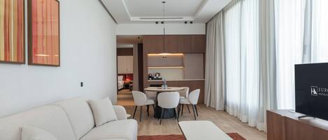 Una habitación de hotel moderna con sofá, una mesa de comedor con sillas, televisión y una zona de cocina al fondo.