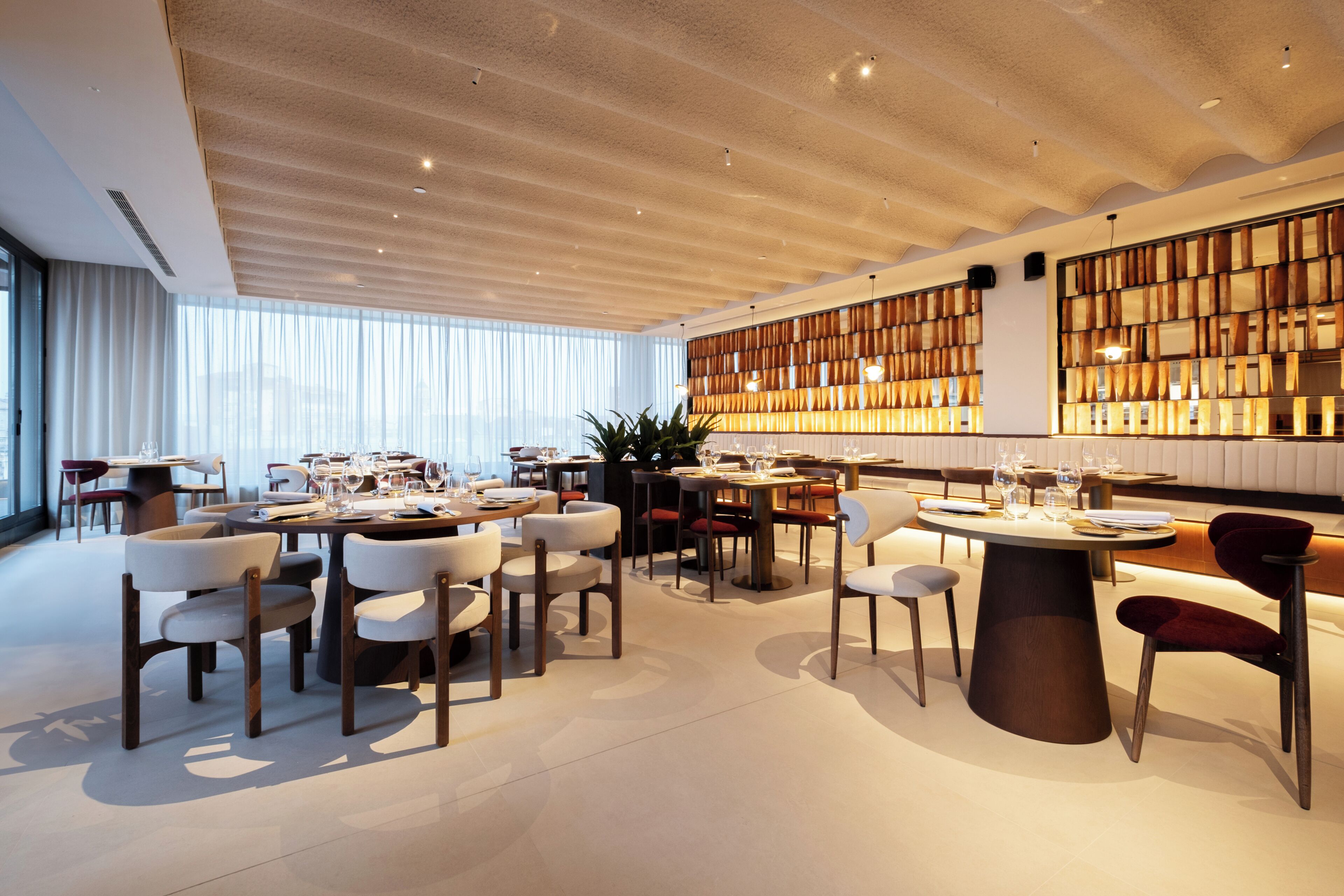 A modern dining area with round tables and chairs, a wooden ceiling, and a large window with curtains.