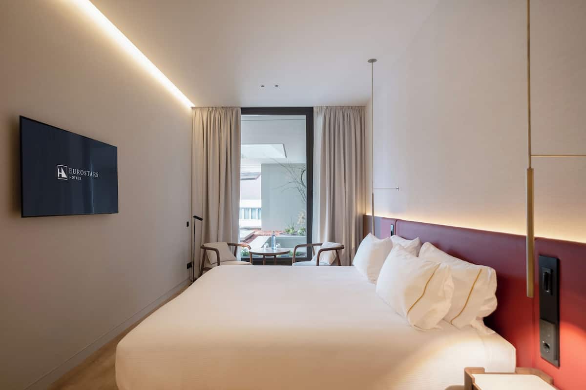 Une chambre d’hôtel avec un grand lit, un téléviseur fixé au mur, une petite table avec des chaises et une fenêtre avec des rideaux.