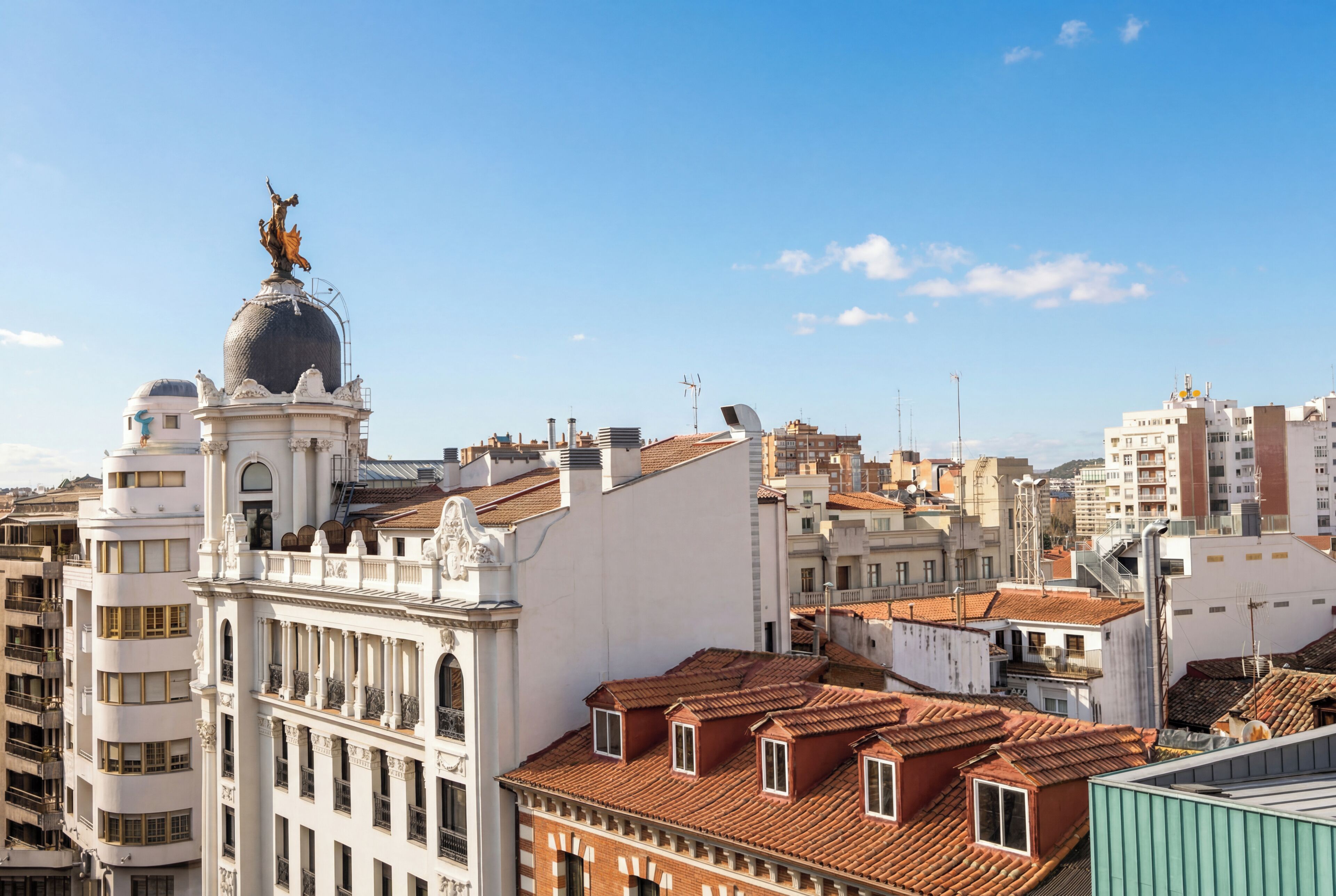 A cityscape with a prominent white building featuring a dome and a statue, surrounded by other buildings with red-tiled roofs.