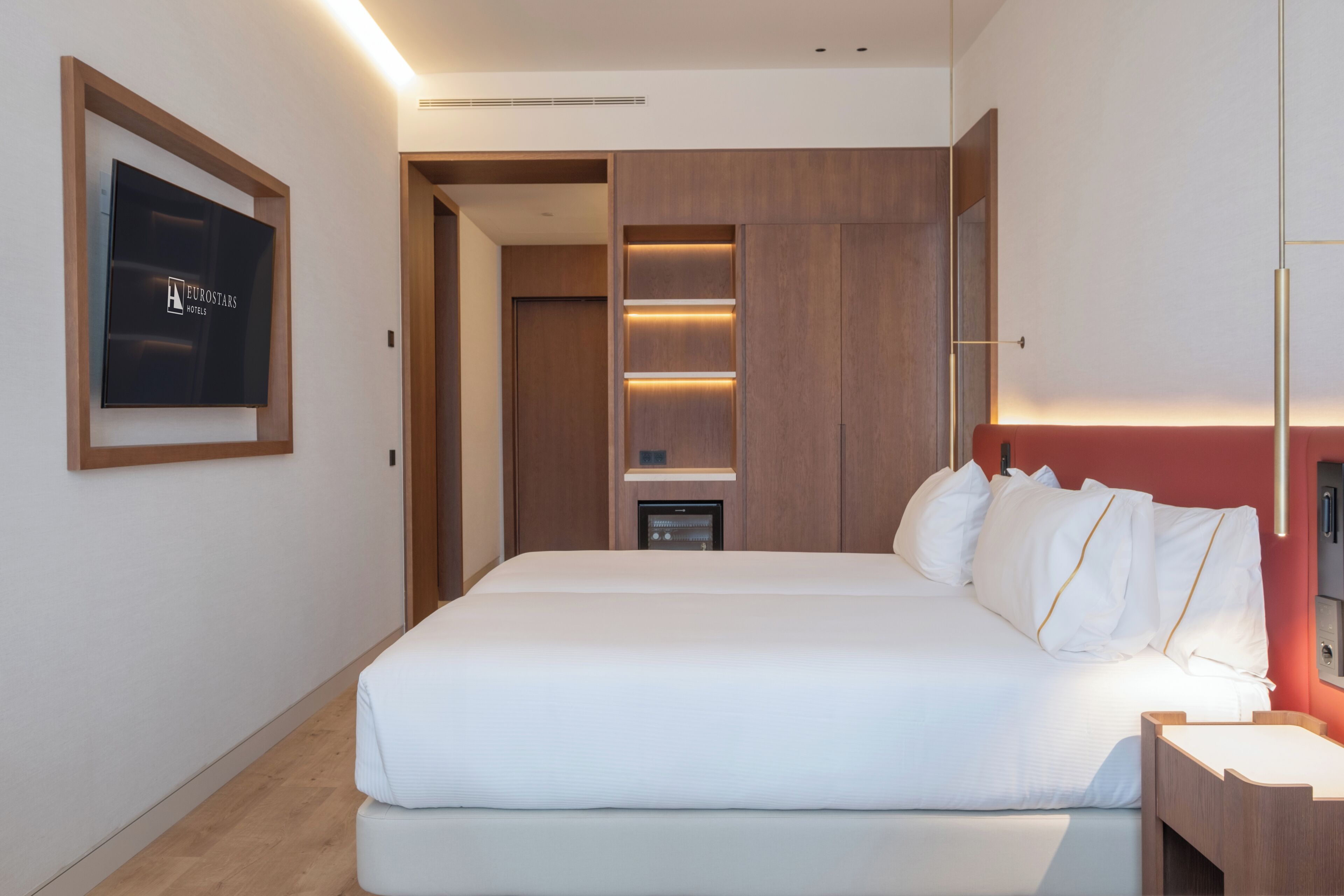 A hotel room with a large bed, a TV mounted on the wall, a wooden wardrobe, and a bedside table.