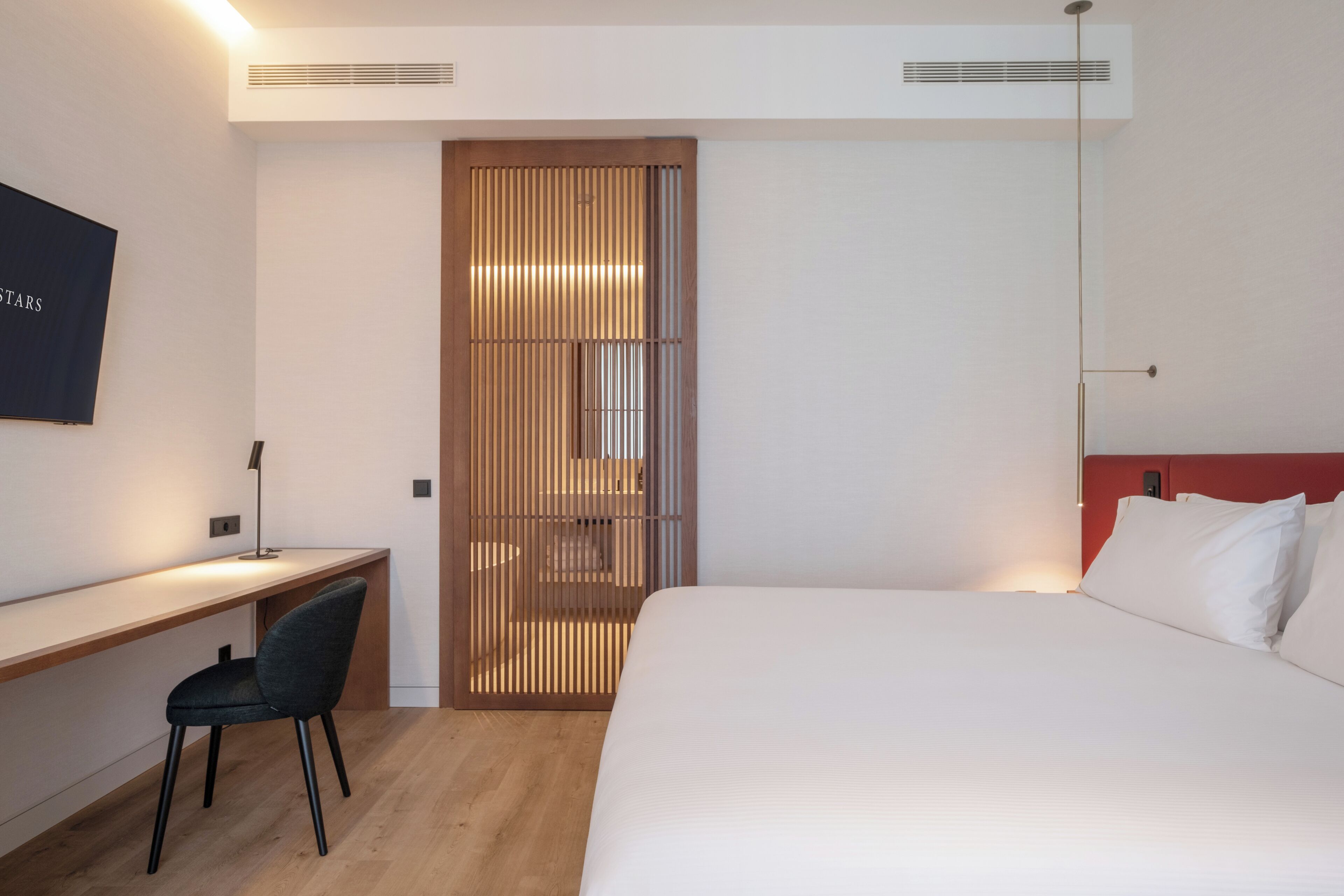 A hotel room with a bed, desk, chair, and a partition with vertical slats.