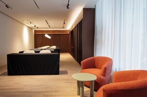A modern hotel room with a long corridor, a bed with a black cover, two orange armchairs, a small round table, and a large window with sheer curtains.