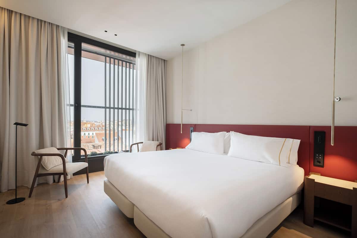 Une chambre d’hôtel avec un grand lit, un bureau, une chaise, une fenêtre avec des rideaux et une vue sur la ville.