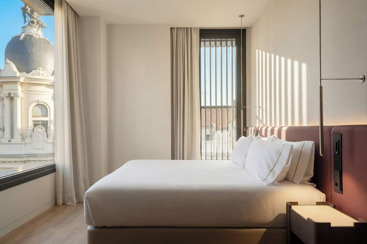 Une chambre d’hôtel avec un grand lit, une fenêtre avec des rideaux, une vue sur un bâtiment doté d’un dôme et un luminaire mural moderne.