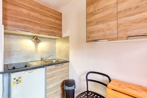Une cuisine compacte avec des meubles en bois, un évier en acier inoxydable, un petit réfrigérateur et une poubelle.