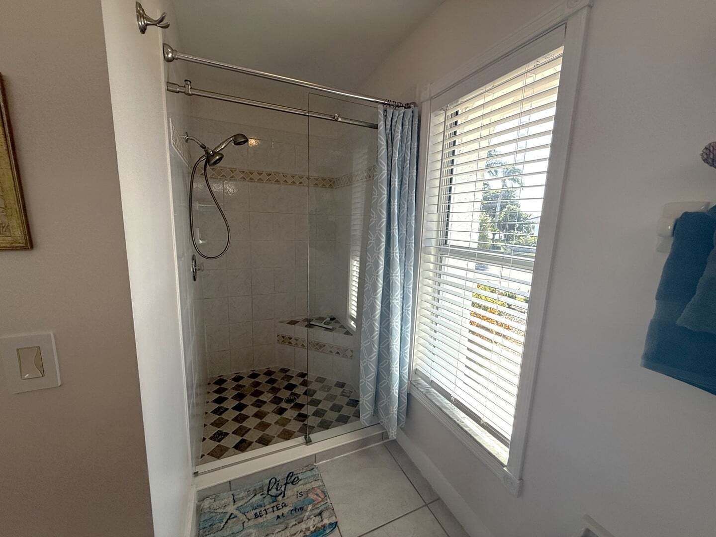 A shower with a glass door, a window with blinds, and a checkered floor mat.